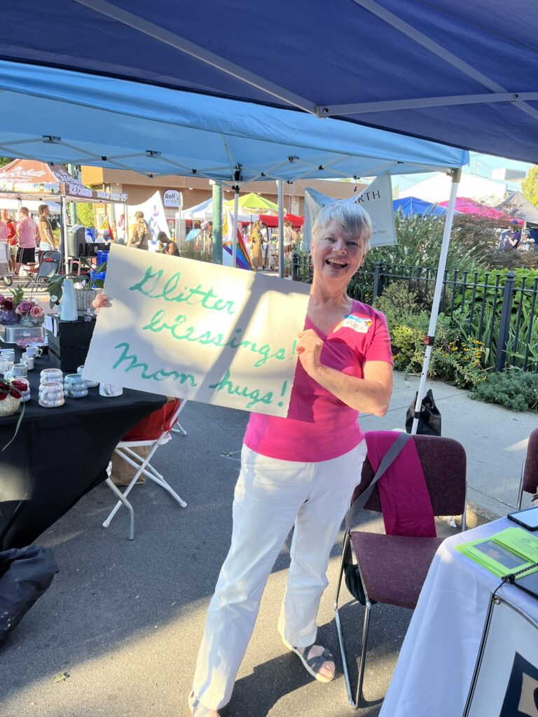 A woman smiles broadly while holding a sign that says "glitter blessings, mom hugs"